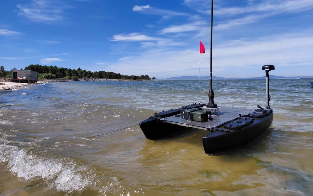 A black catamaran unmanned surface vessel with a tall antenna mast in shallow water by a sandy shore